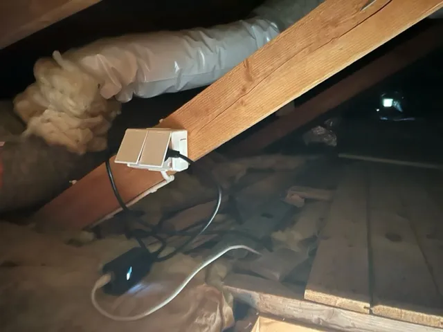 Isolerade ventilationskanaler och en grå flexslang vid en takbjälke på vinden, med ett eluttag på bjälken och en nätadapter på isoleringen